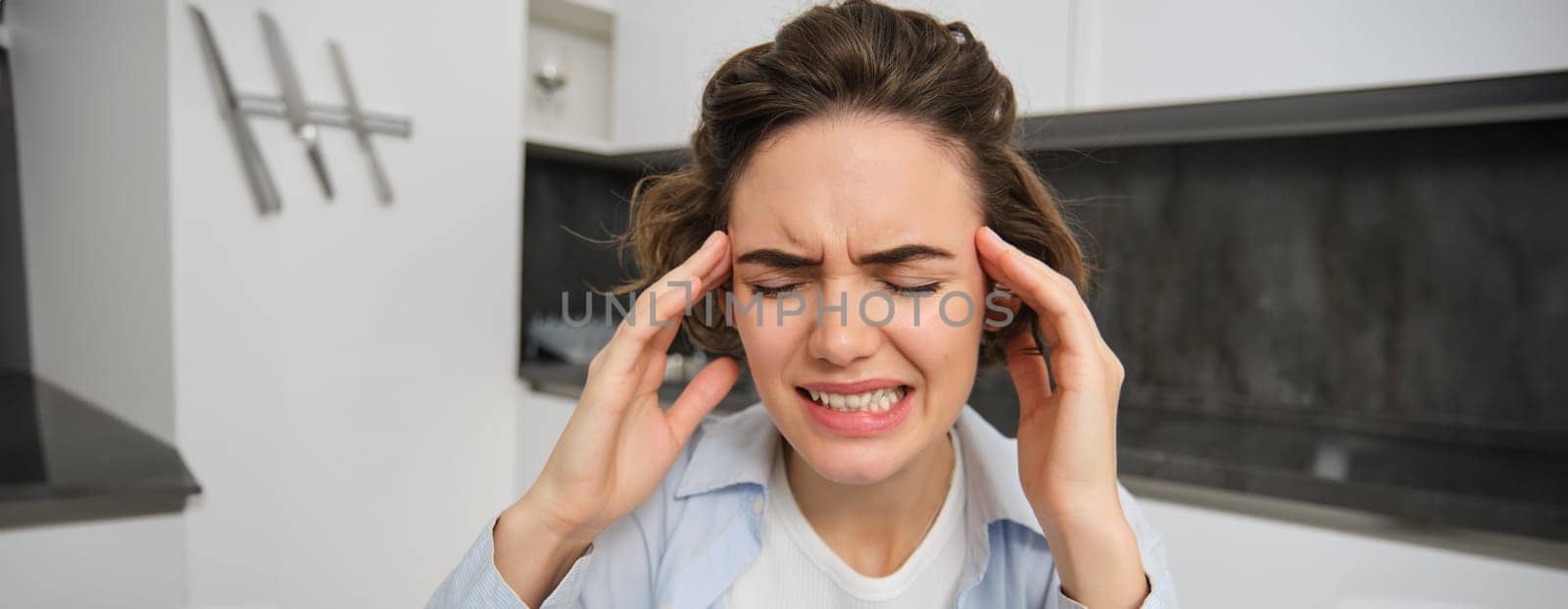 Portrait of woman with headache, sits in kitchen, touches head and grimaces, has painful migraine by Benzoix