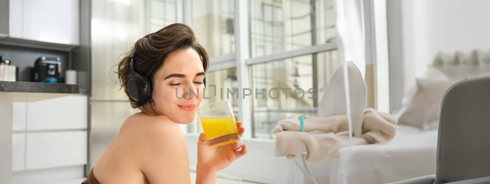 Smiling sportswoman, fitness girl does workout at home, wears headphones, listens music and drinks orange juice after training session by Benzoix