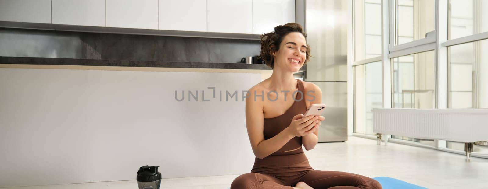 Sport and women lifestyle. Young woman does workout at home, sits on yoga mat with smartphone in bright kitchen, wears activewear, uses mobile phone app for indoor exercises by Benzoix