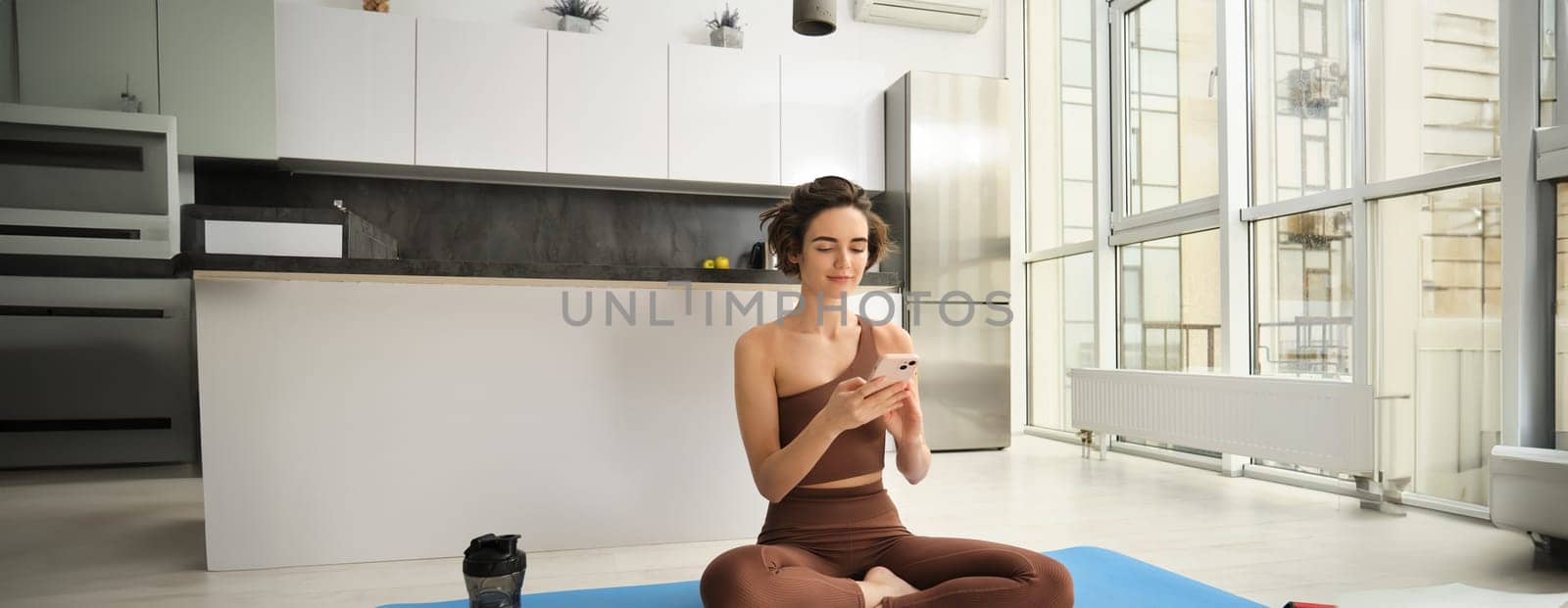 Yoga and sport at home concept. Young woman sitting in bright rome with mobile phone, doing workout on rubber mat, searching for exercises online on smartphone app, using sports equipment.