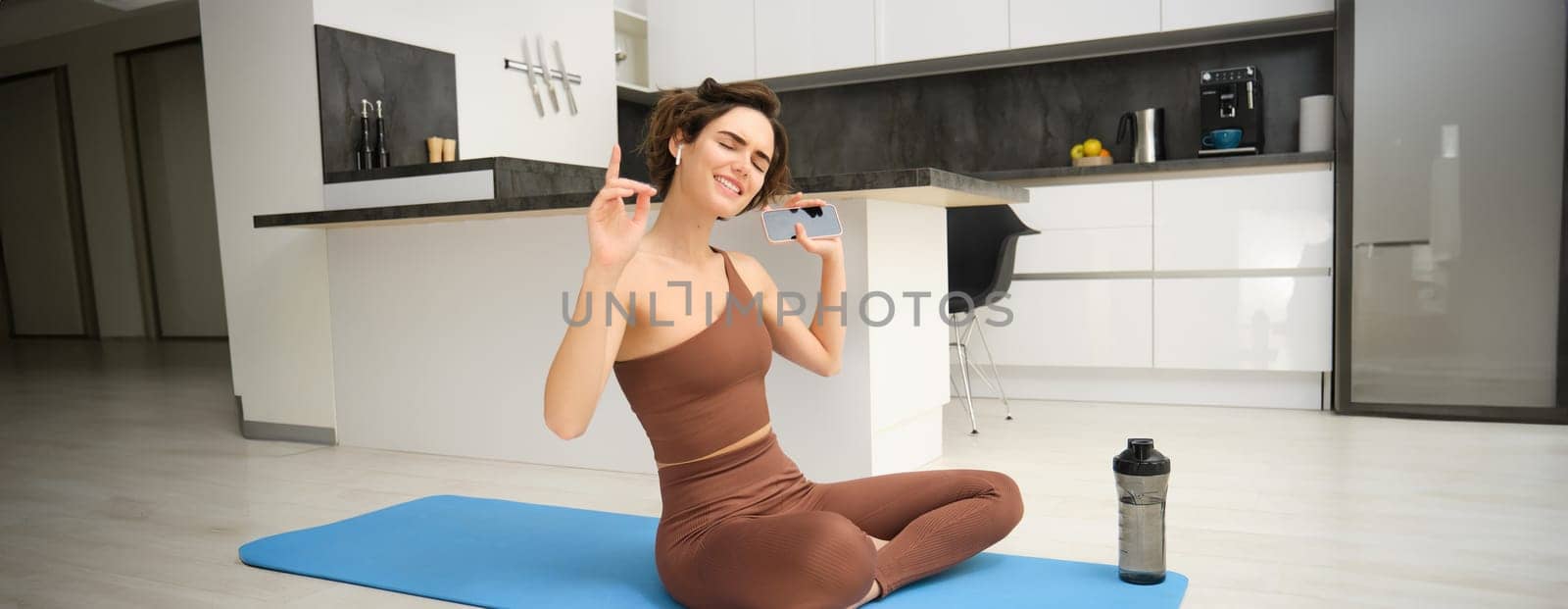 Active lifestyle and health. Happy young woman does workout, sits and dances with smartphone in hands, listens music in wireless earphones during fitness training session, yoga on floor mat by Benzoix