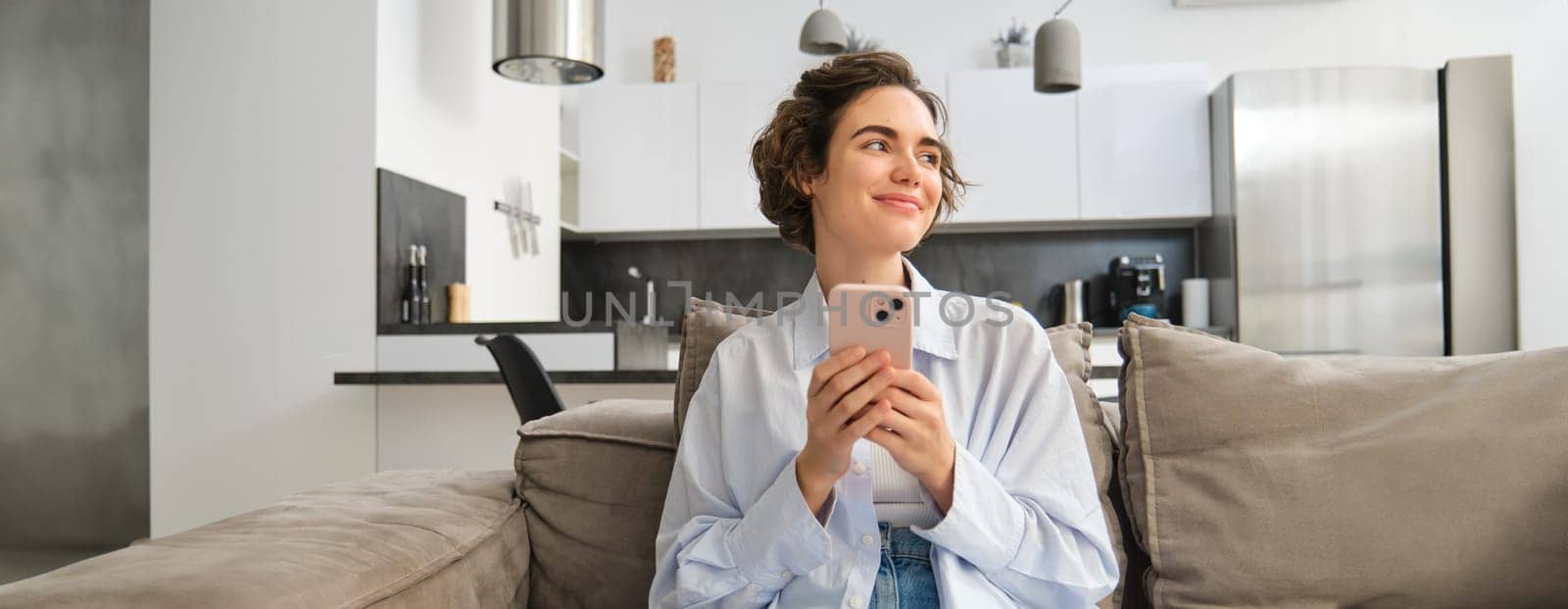 Image of brunette smiling woman with smartphone, sitting at home on sofa, looking aside with happy, thoughtful face, chatting on phone, using mobile app.