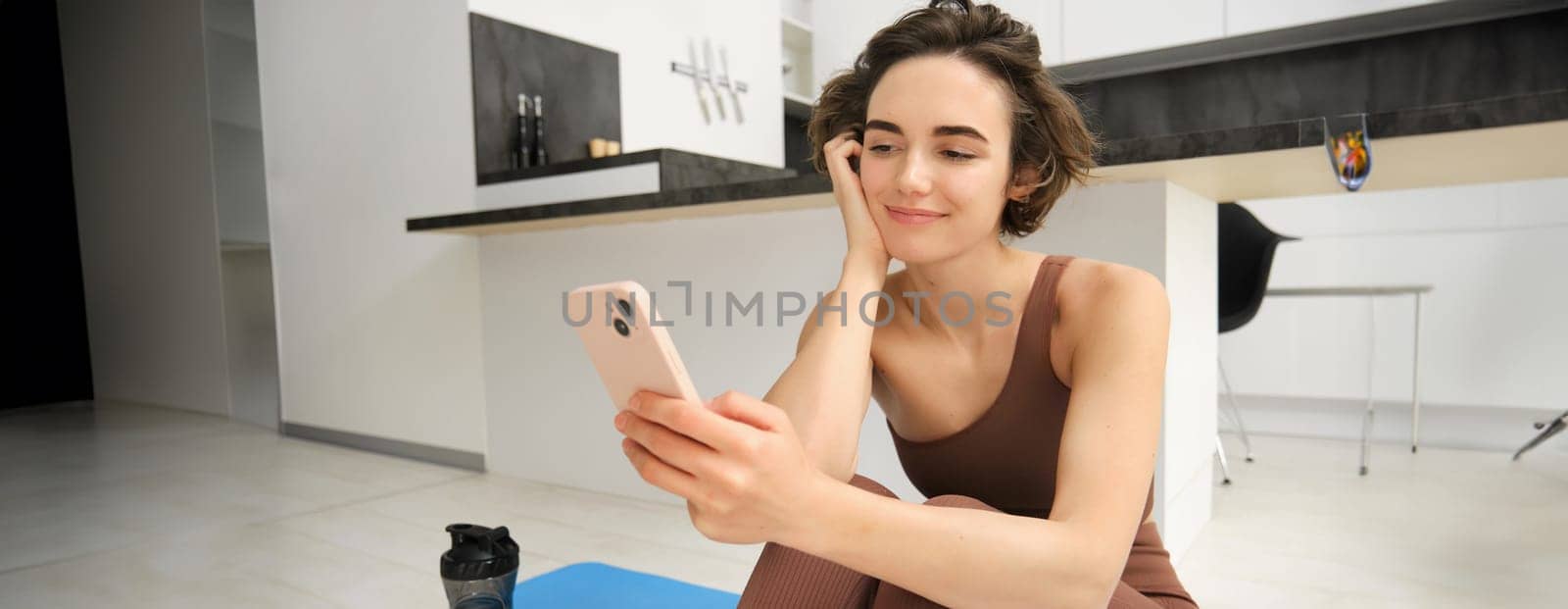 Technology and sport concept. Fitness girl looking at her smartphone, sitting in workout clothes at home, watching exercise videos on mobile phone.