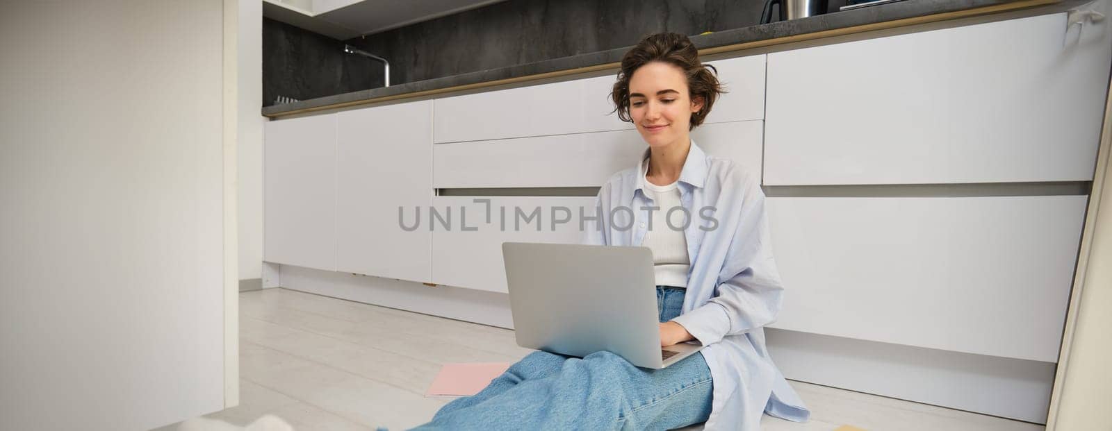 Portrait of girl freelancer, sits at home on floor and works with laptop, surrounded with piles of documents, smiles while looks at computer screen.