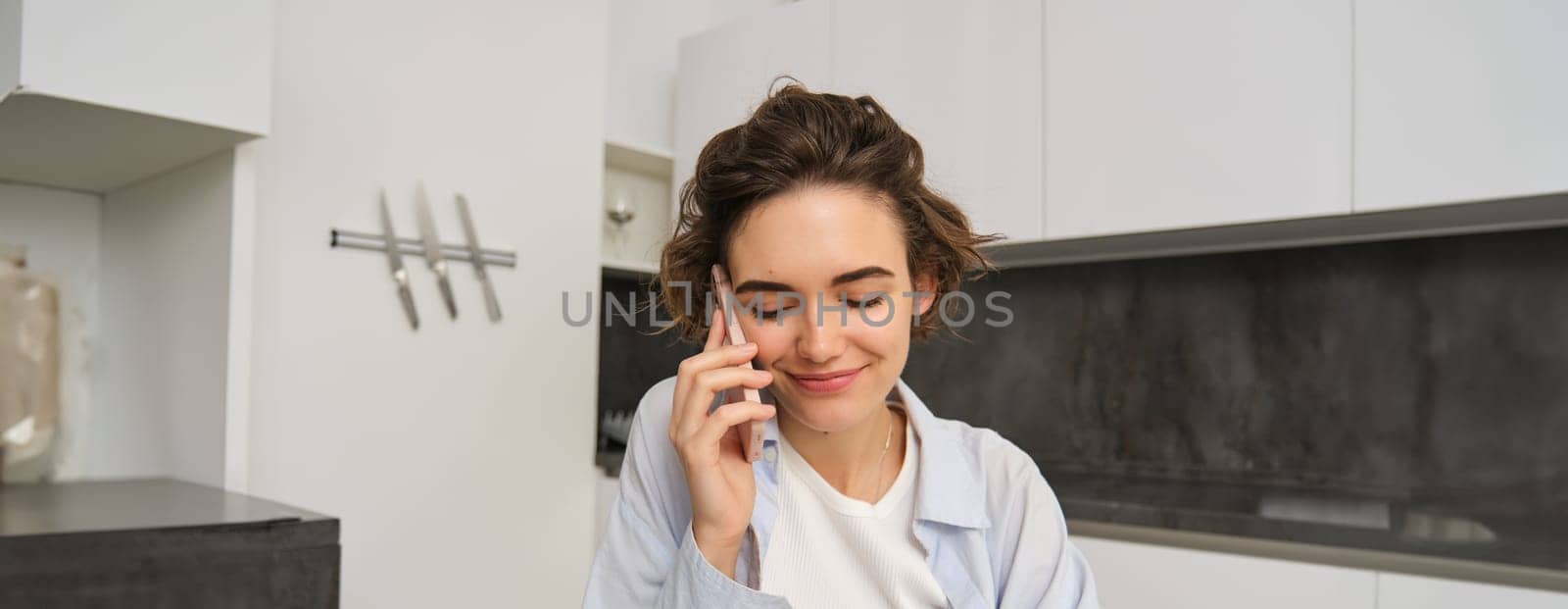 Portrait of beautiful girl talks on mobile phone, has conversation, answer a call in kitchen by Benzoix