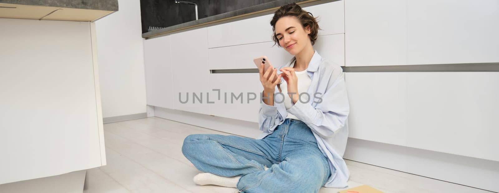 Portrait of cute smiling woman, sitting on floor at home with crossed legs, looks at mobile phone, chats on smartphone app, watches videos in application.