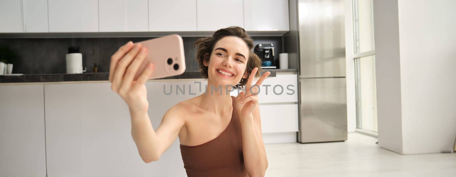 Smiling fitness girl, takes selfie during workout at home, posing for photo and holding smartphone, workout from home, recording vlog of her yoga training session by Benzoix