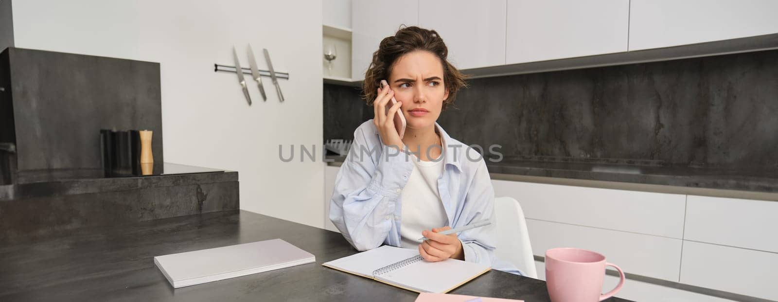 Portrait of woman working from home, calling someone on mobile phone, looking confused while holding pen and trying to write down something, sitting with puzzled face in kitchen by Benzoix