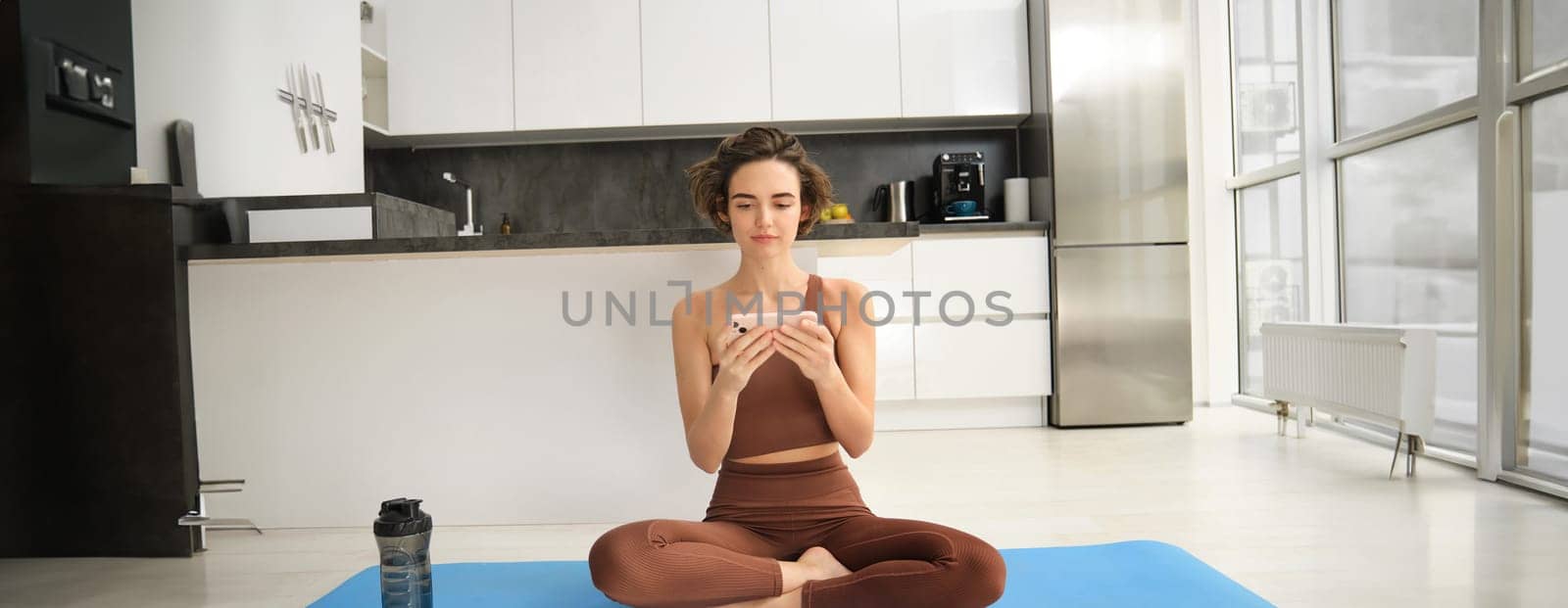 Woman sits on yoga rubber mat, watching fitness video tutorial on smartphone app, making lotus pose, workout at home, learns pilates indoors. Sport and wellbeing concept