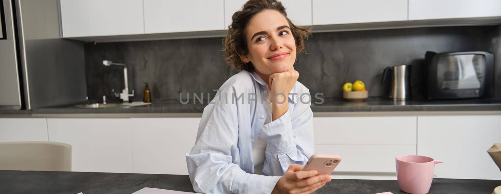 Beautiful, dreamy young woman, holds smartphone, smiles and looks thoughtful, orders takeaway, groceries delivery on mobile app.