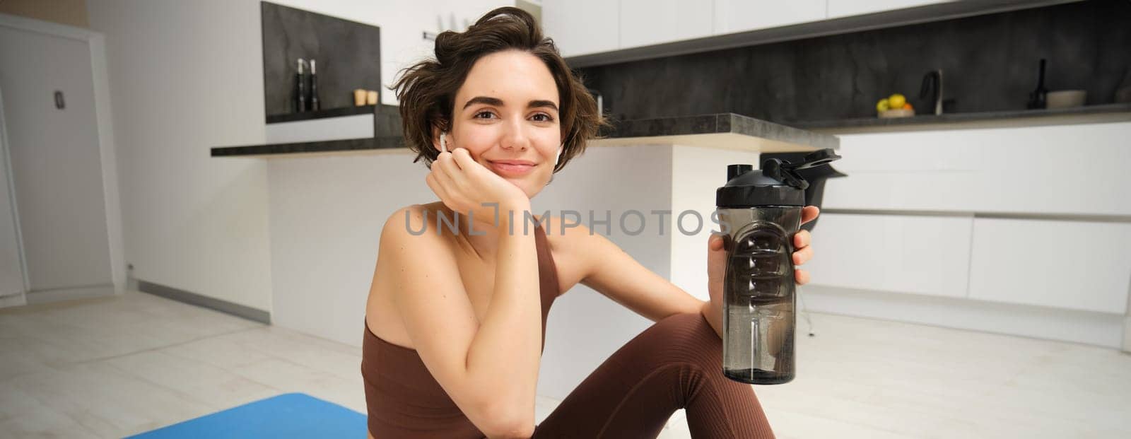 Portrait of fitness woman, young sportswoman at home, drinks water from bottle after workout, training exercises, takes a break after pilates, yoga training, stays hydrated.