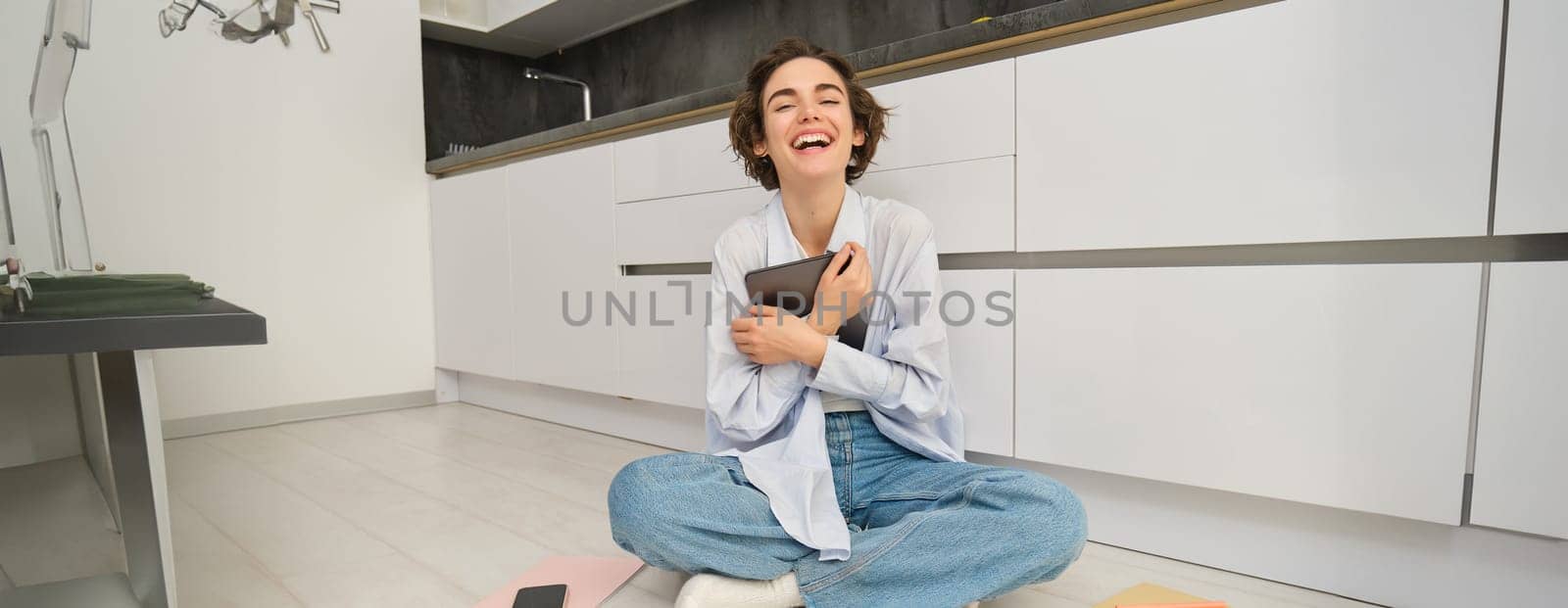 Portrait of happy woman with tablet, sitting on floor, laughing and smiling, working from home on remote, studying online by Benzoix