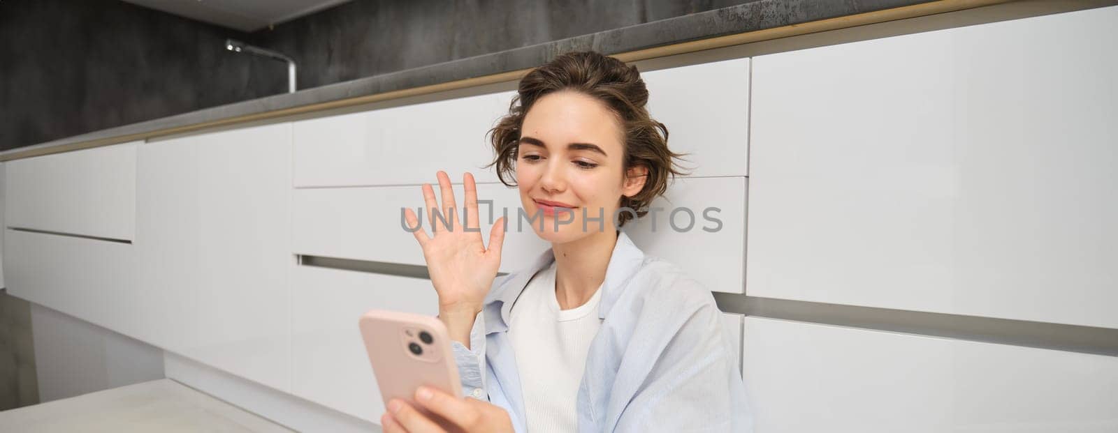 Happy girl sits on floors and video chats on smartphone app, connects to conversation on her mobile, waves hand at phone camera.