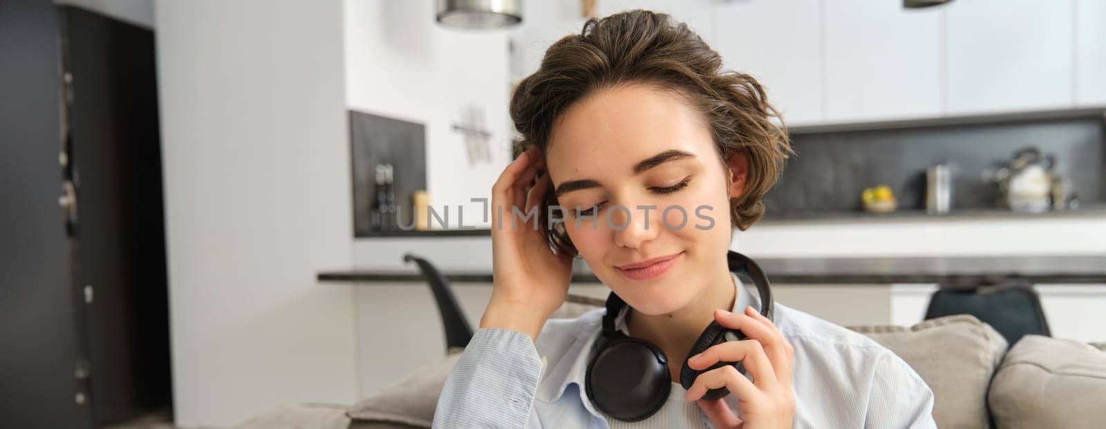 Close up portrait of woman enjoying listening to music in headphones, sitting at home on sofa with earphones on head.