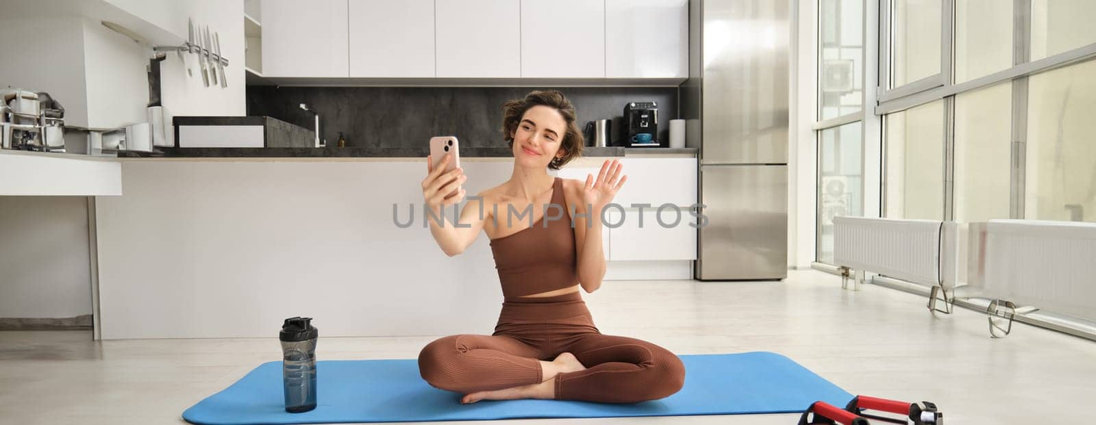 Portrait of young fitness woman, sitting at home on rubber mat, records video on smartphone, doing workout vlog, making live online training session from home by Benzoix