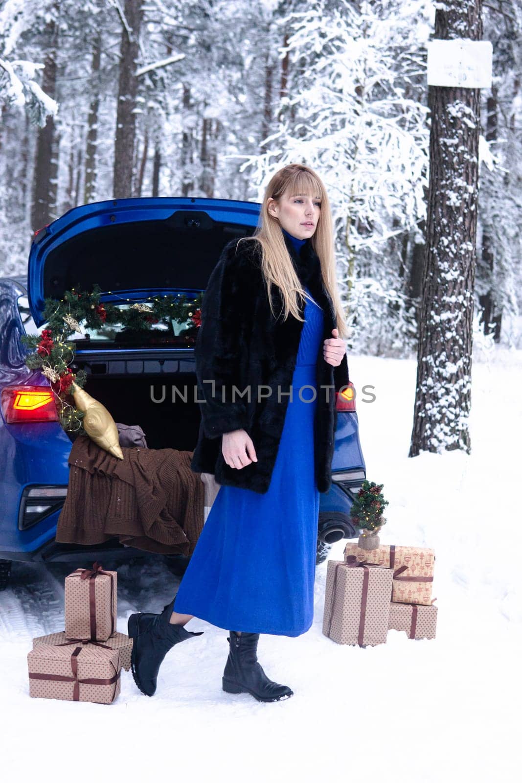 Woman in winter snowy forest in blue dress next to blue car decorated with Christmas decor. Christmas and winter holidays concept. by Annu1tochka