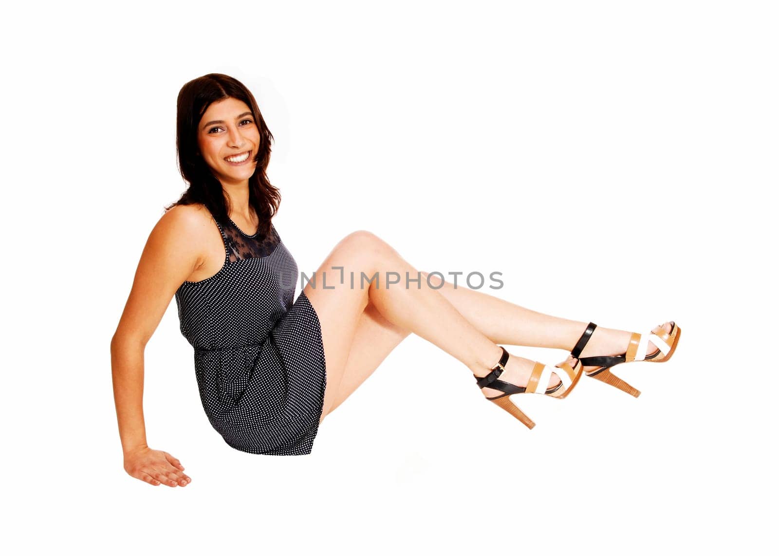 A smiling young woman in a dress sitting in profile on the floor with her.long legs, isolated on white background..