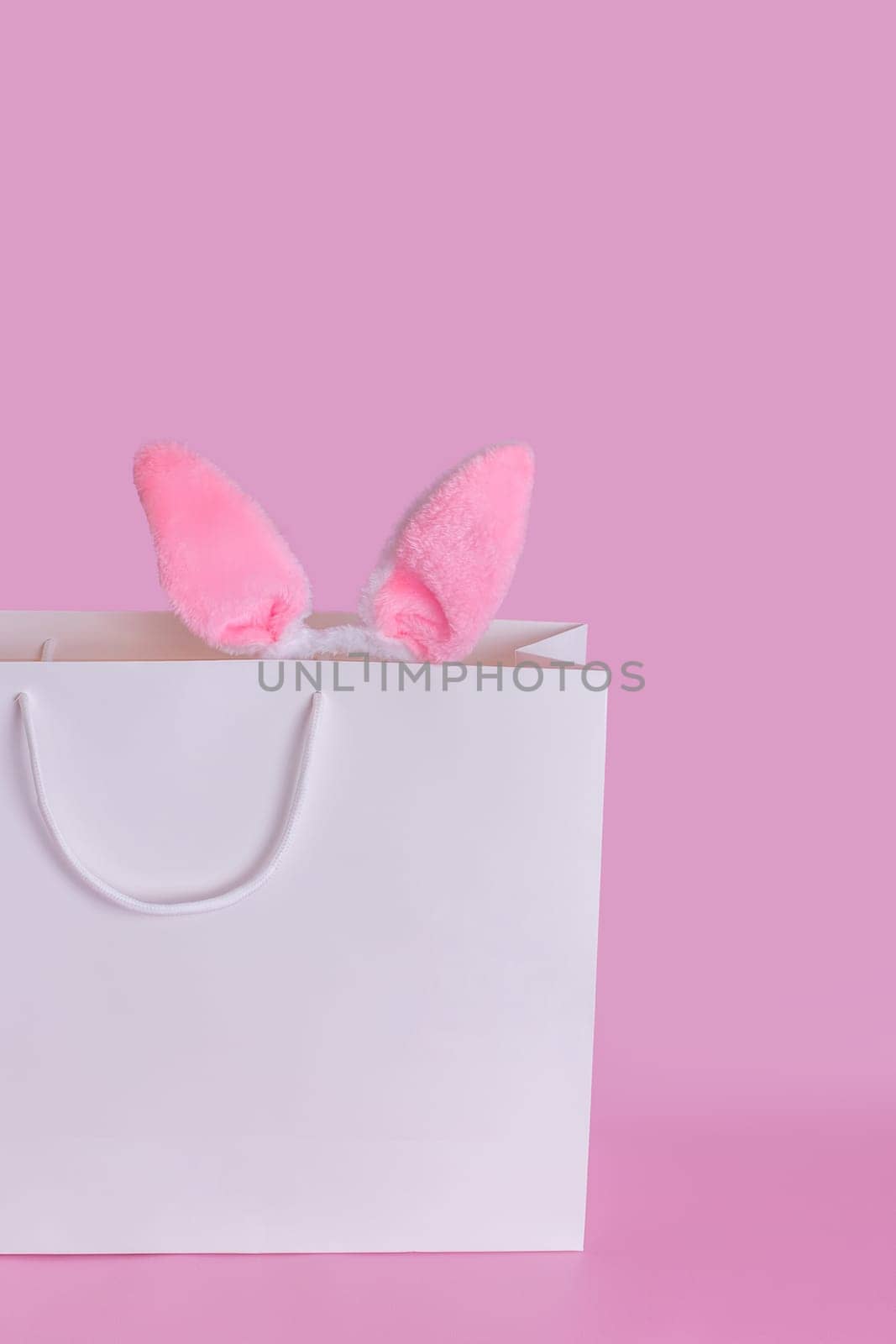 The ears of the Easter bunny in a large white paper gift bag standing on a pink background. Space for text. Vertical