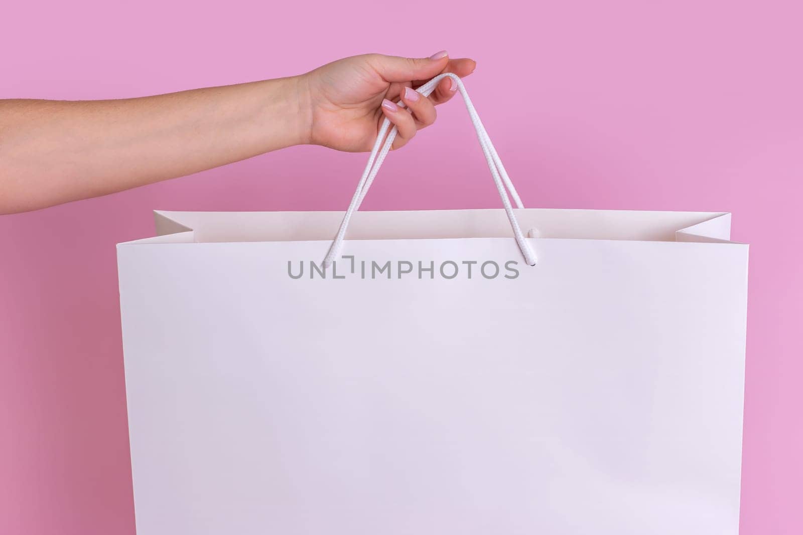 A close-up of a womans hand holding a large white transparent gift bag made of kraft paper for shopping after shopping, highlighted on a pastel pink background. layout. Advertising layout of the delivery concept