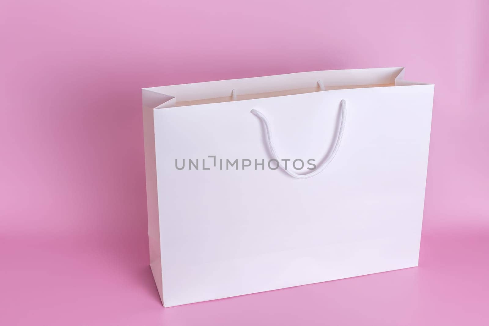A large white paper bag with white rope handles stands on a pink background in close-up. Copy space. Mock up. Top view