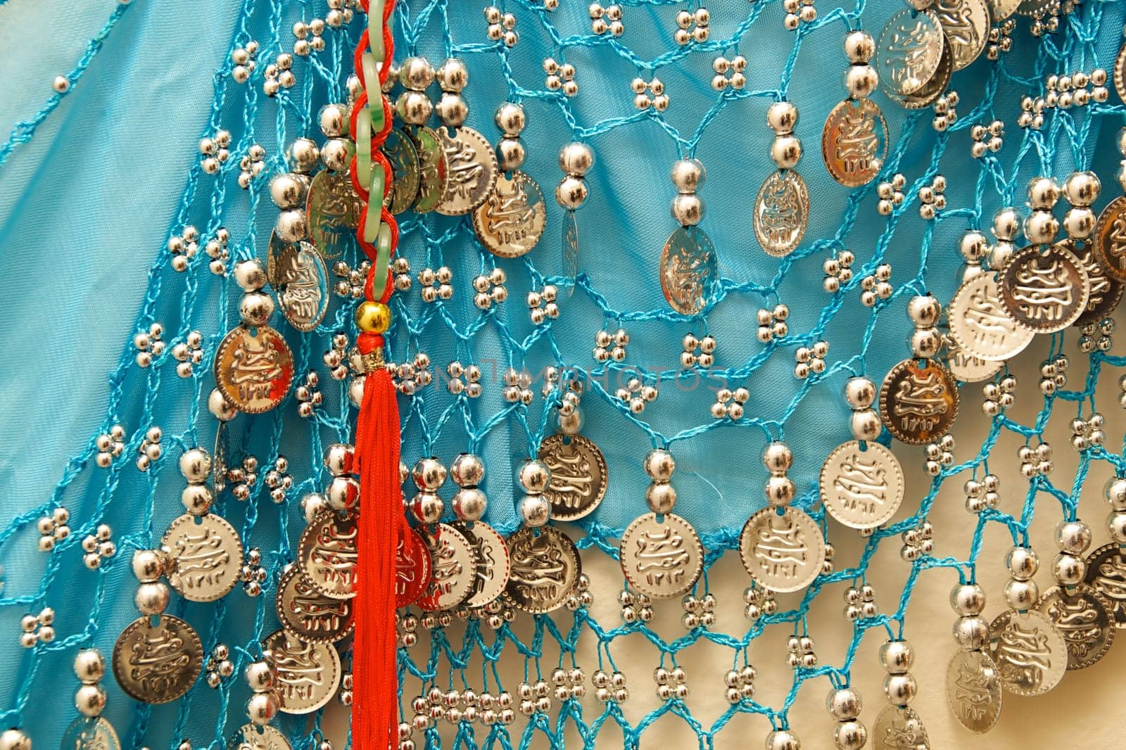 A blue vintage belly dancer's skirt hanging on wall showing silvery beads and charms with red tassel.