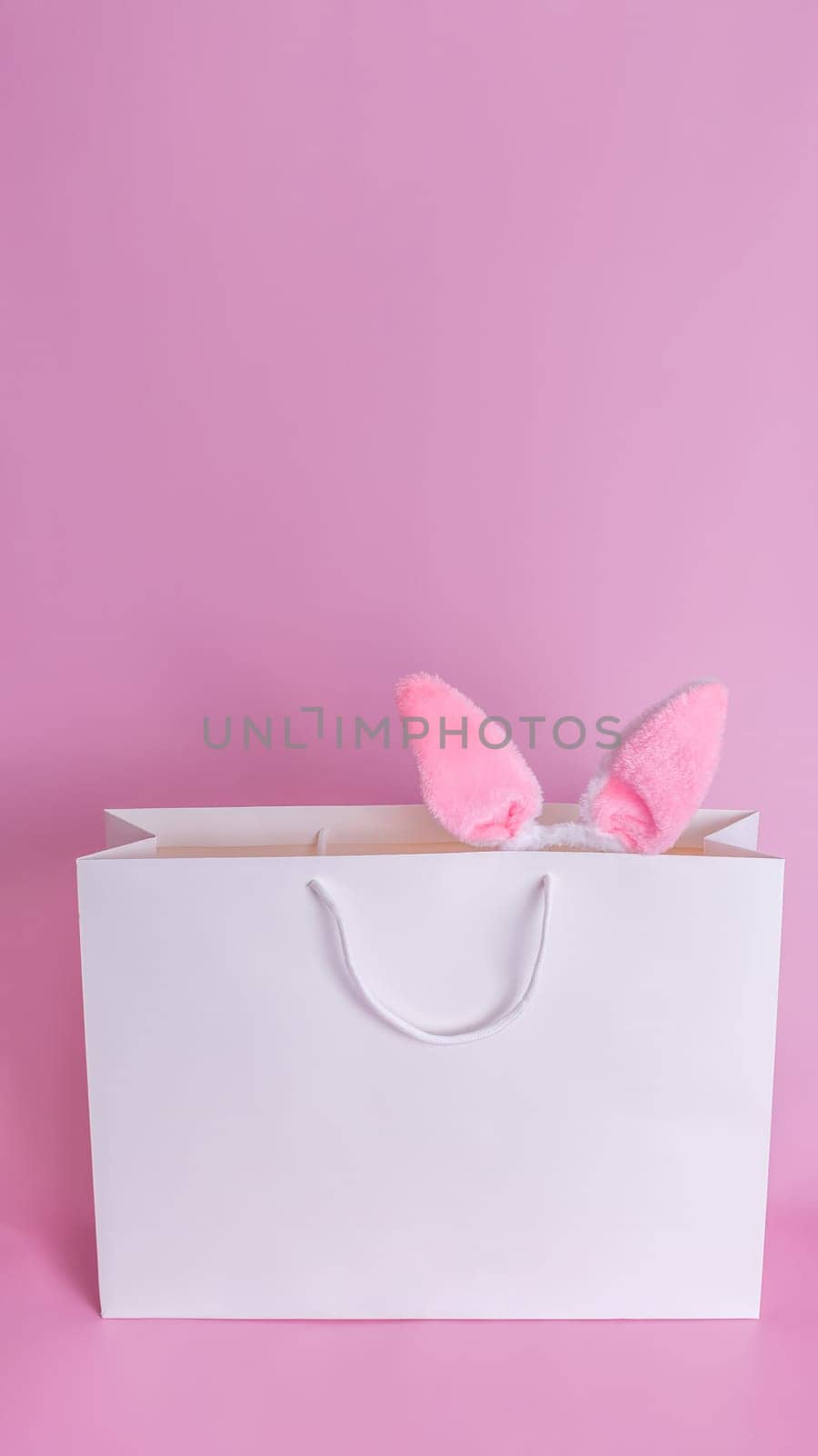 The ears of the Easter bunny stick out of a large white paper gift bag standing on a pink background.Space for text. Happy Easter concept