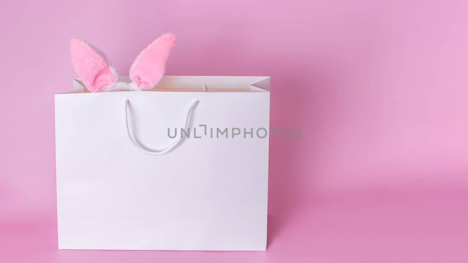 Funny pink ears of the Easter bunny stick out of a large white paper gift bag standing on a pink background. Copy space. Happy Easter concept