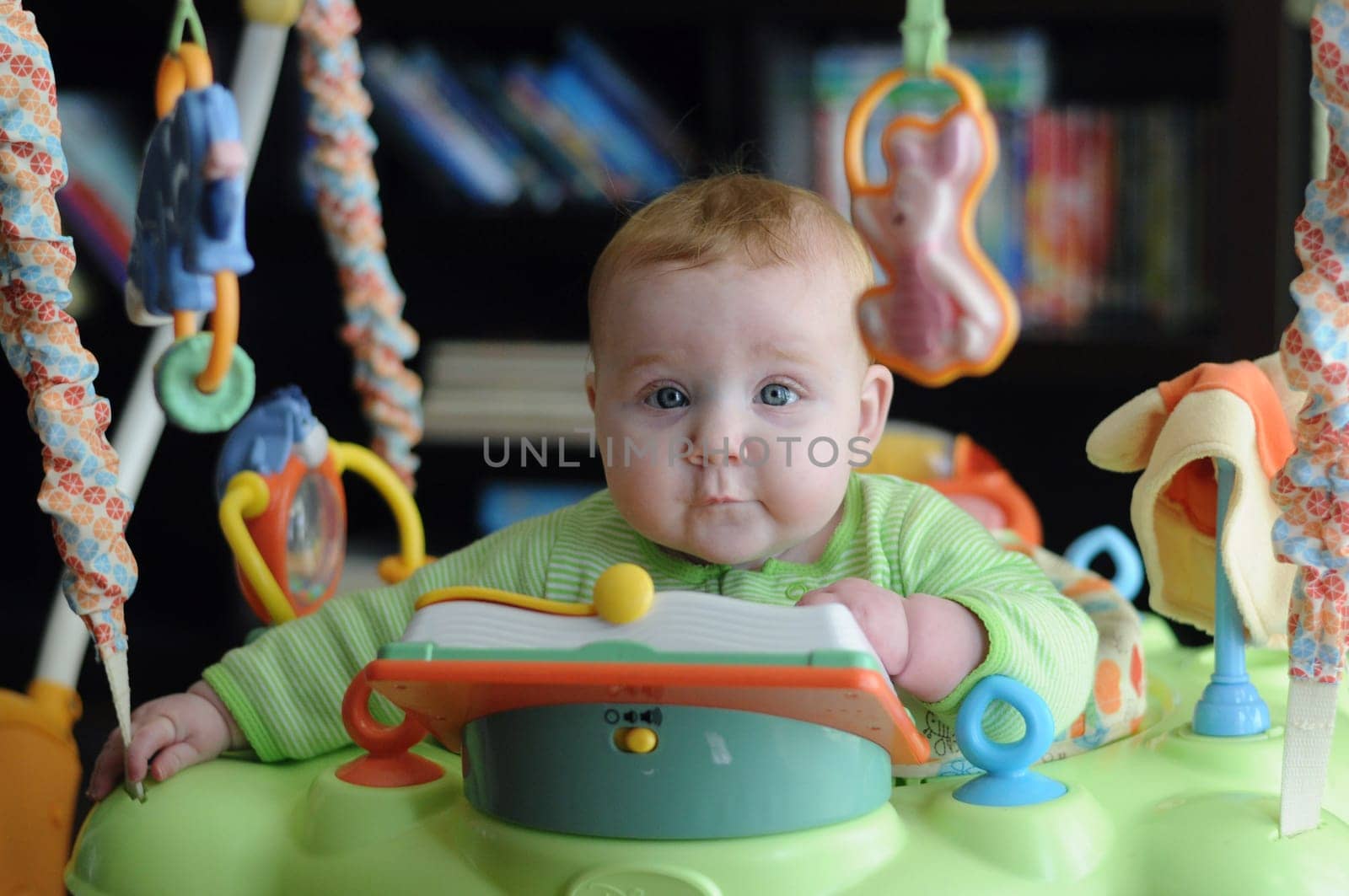 Editorial cute attentive intelligent young baby surrounded by toys in a green outfit sitting looking quizzically at the camera