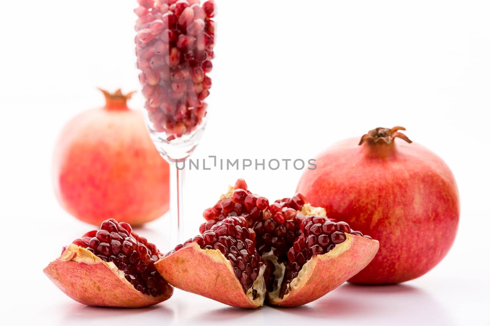 Ripe, deep red pomegranate seeds, embedded in its reddish fruit skin. Separated seeds in a slim champagne glass. Two whole pomegranate fruits..