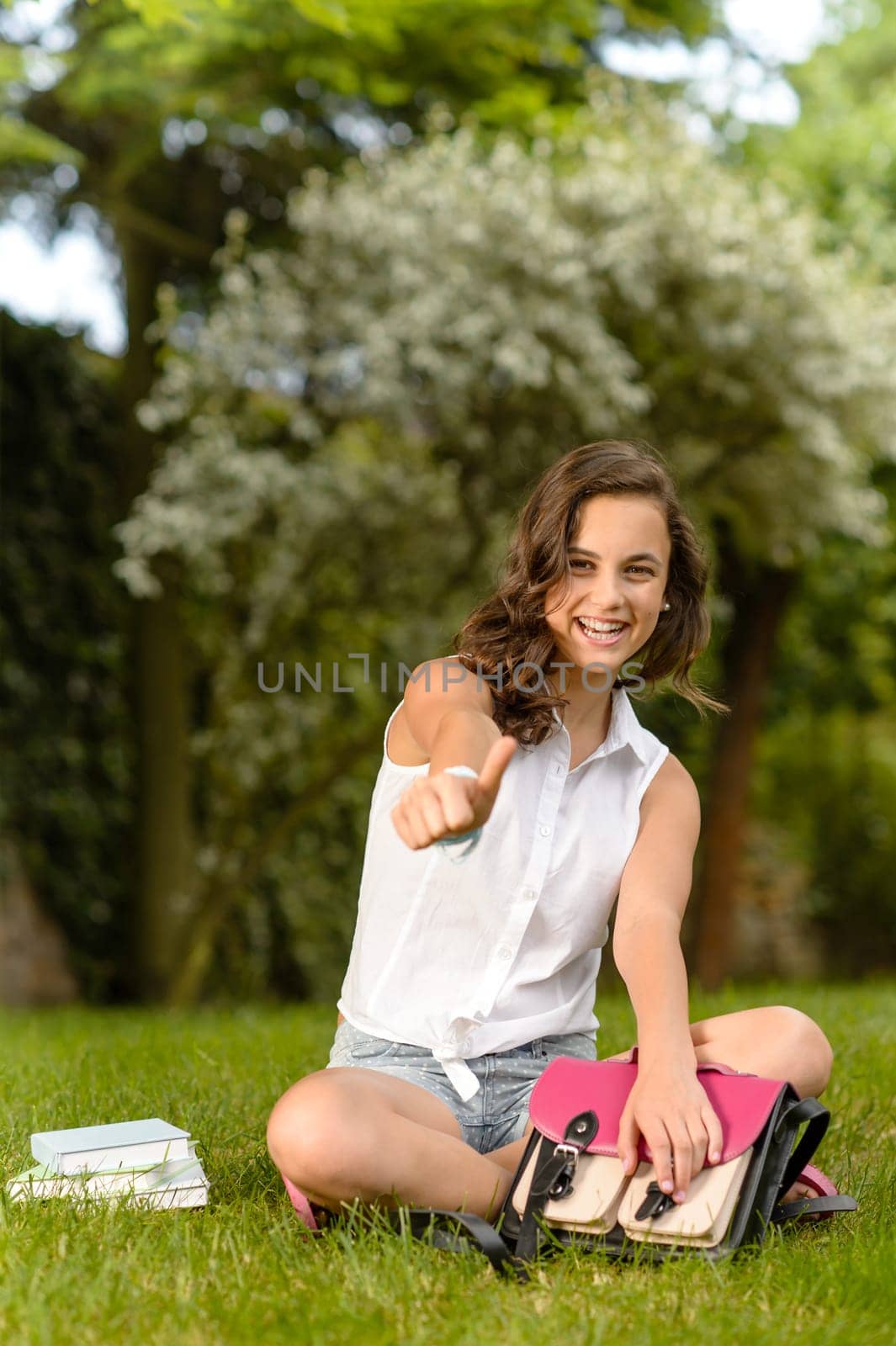 Cheerful student girl sitting on grass shows thumb up summer
