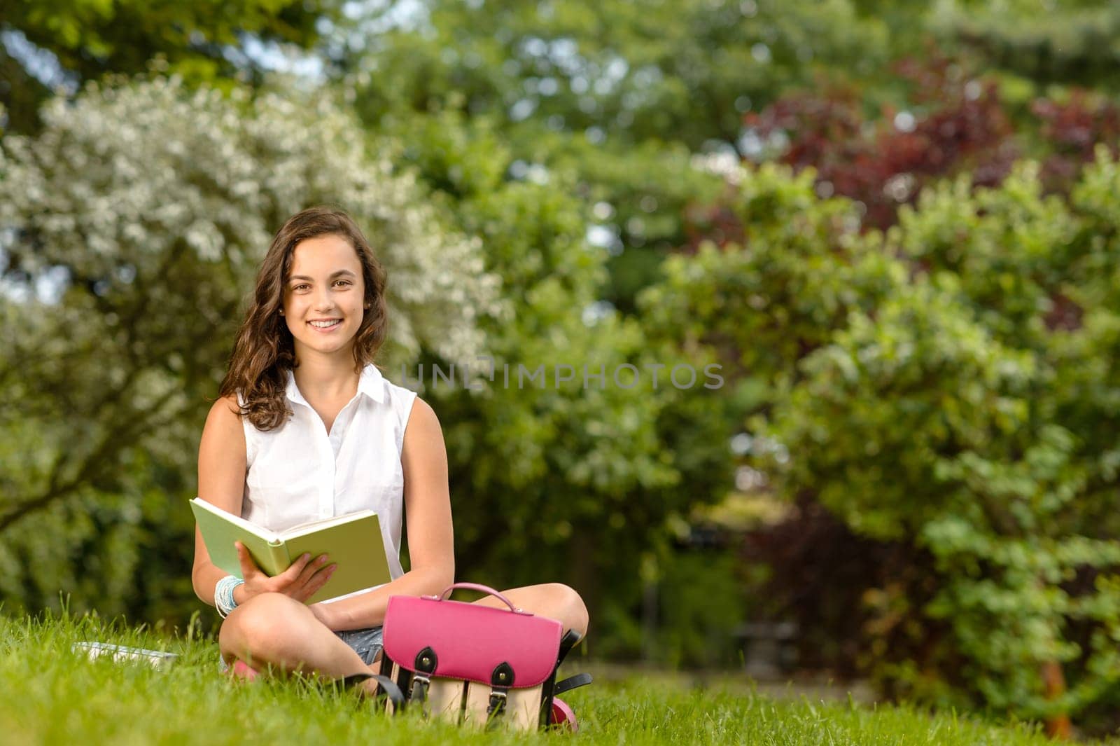 Smiling student girl sitting on grass with open book summer