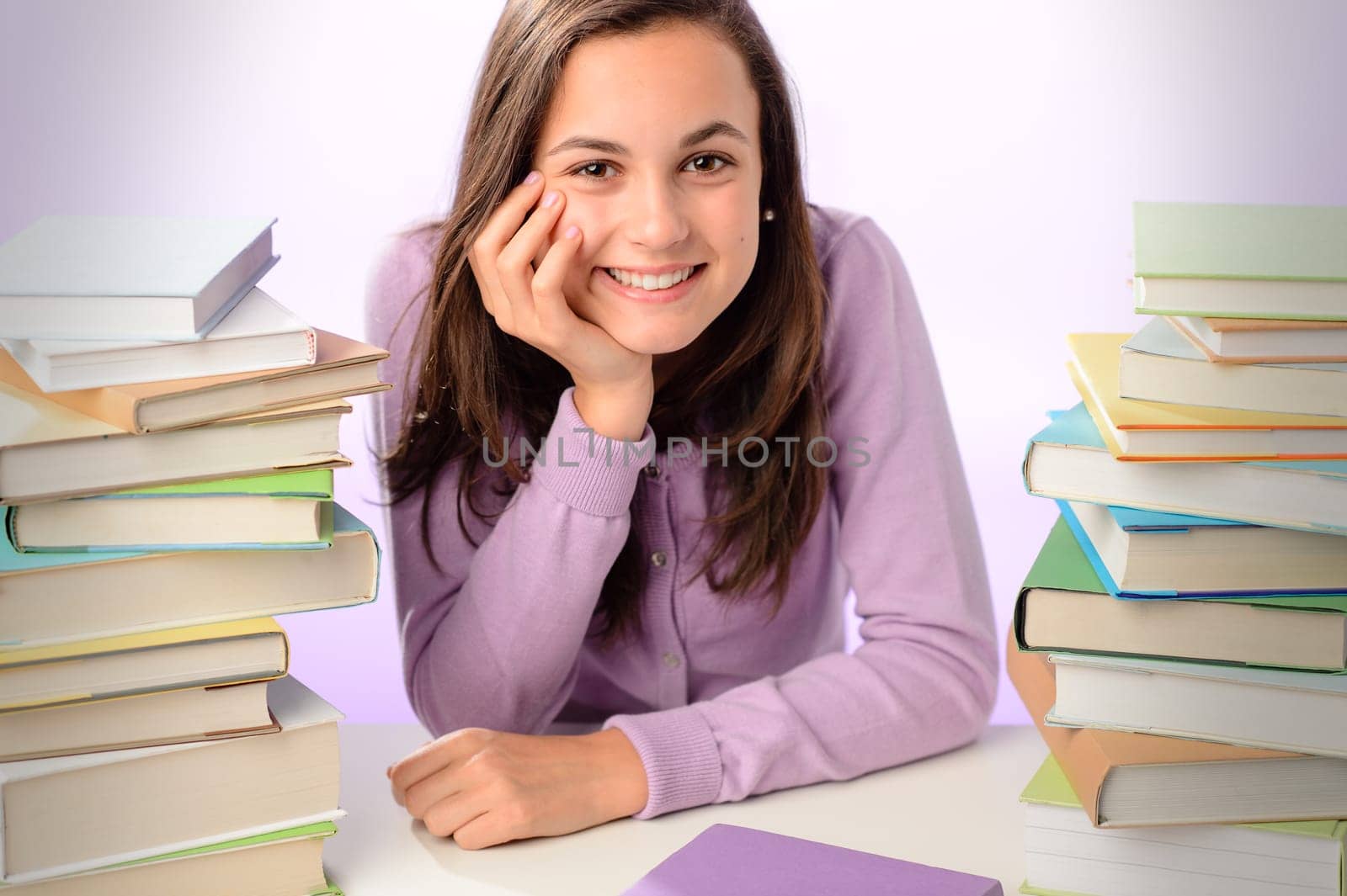 Smiling student girl sitting between stacks of books purple background