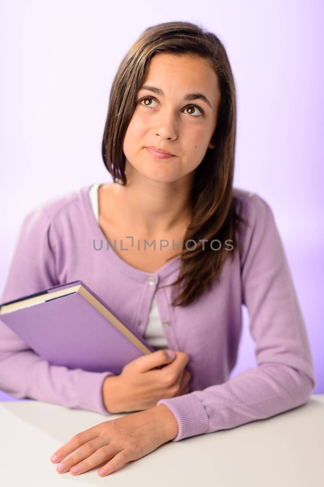 Thinking student girl sitting behind desk holding book purple background