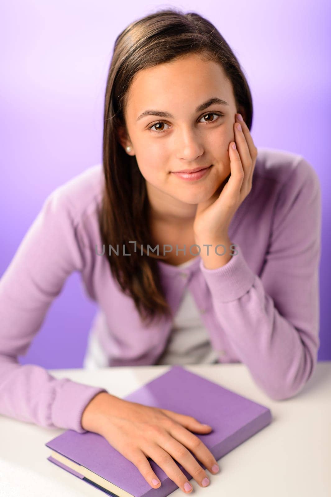 Cute student girl with book sitting behind desk purple background