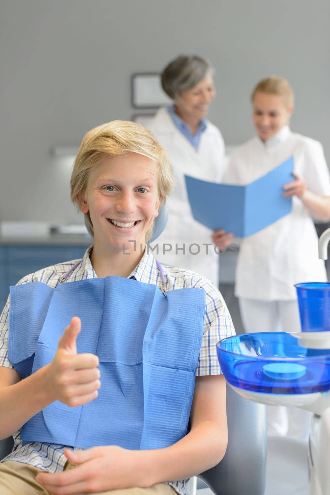 Teenager patient sitting at dental surgery thumbup dentist assistant background