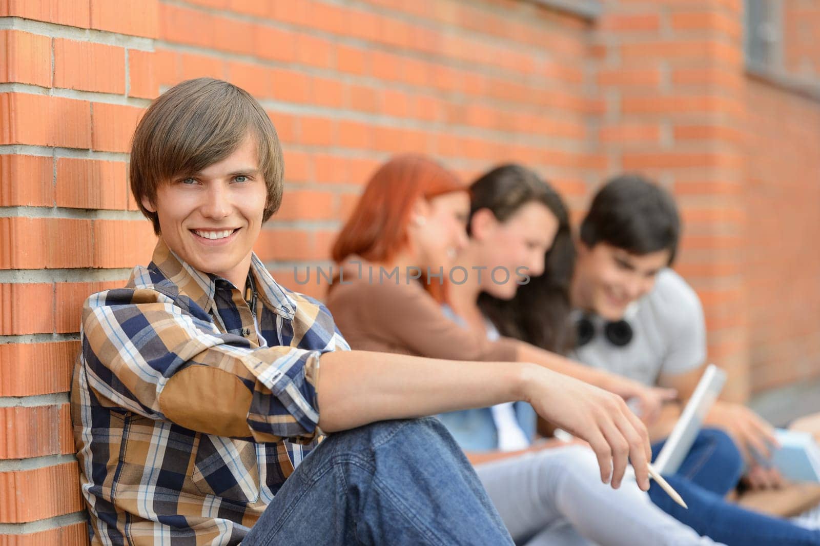Student boy sitting by brick wall friends smiling in background