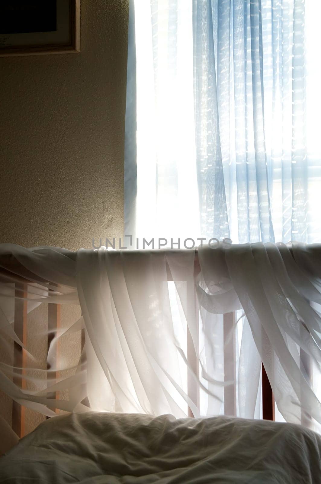 A simple bedroom under ambient light from the window coming in through blue sheer curtains to light up the sheer fabric wrapped around the headboard of the bed.