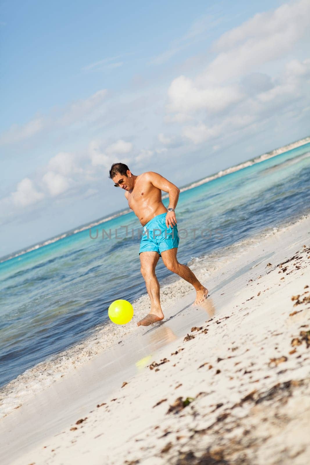 Young attractive man in swim shorts plays football on the beach on holiday in summer