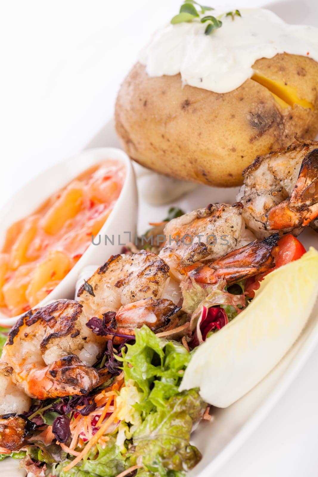 Roasted shrimp on a skewer on mixed salad with dip and foil potato prepared on a plate insulated against a white background