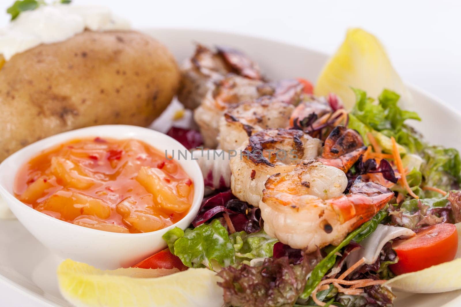 Roasted shrimp on a skewer on mixed salad with dip and foil potato prepared on a plate insulated against a white background