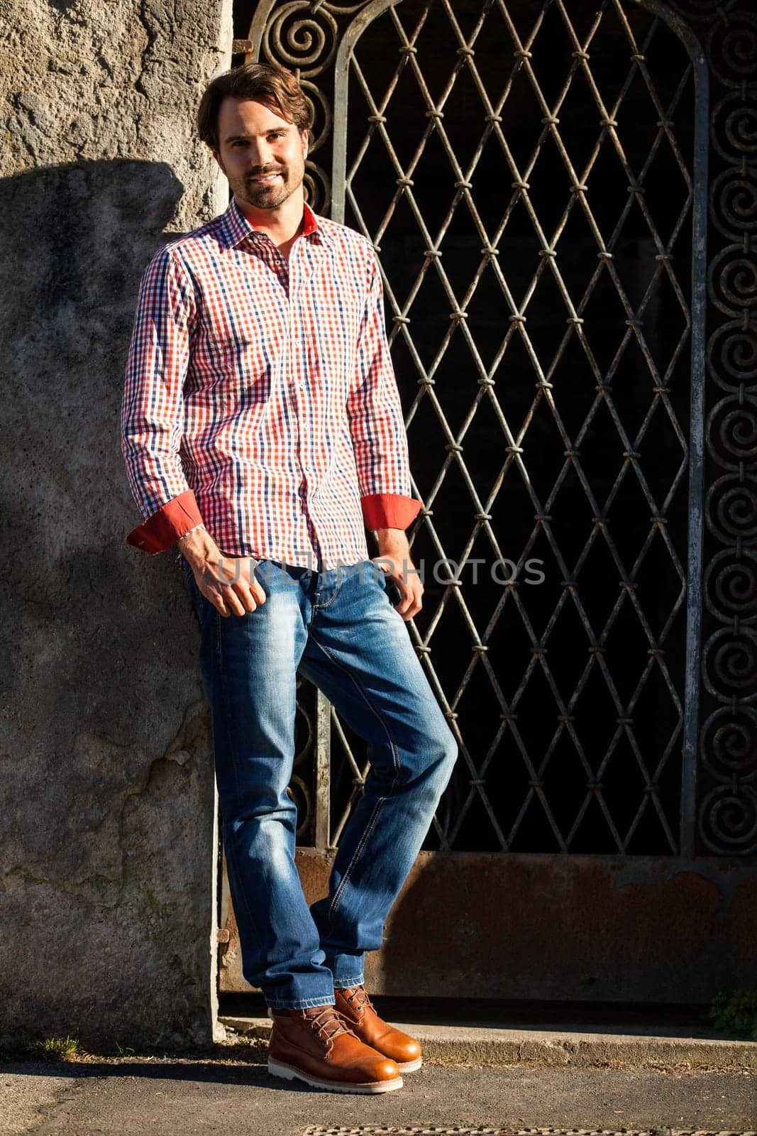 Attractive young man with a plaid shirt and jeans leaning against a wall outdoors in summer
