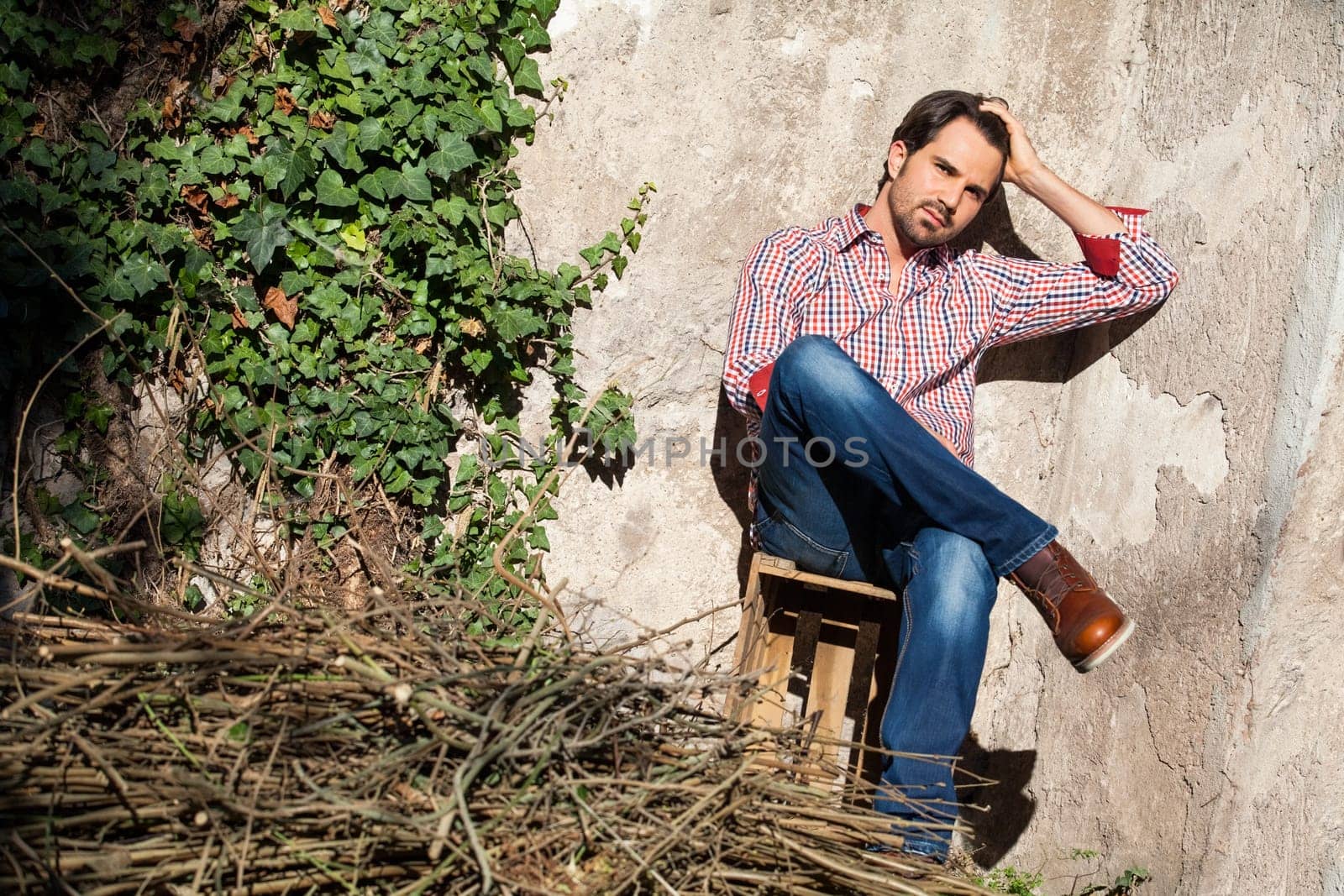 Young man with a plaid shirt and jeans with dark hair and beard outdoors under an efeuranke in daylight