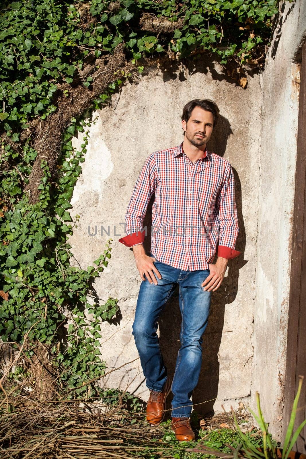 Young man with a plaid shirt and jeans with dark hair and beard outdoors under an efeuranke in daylight