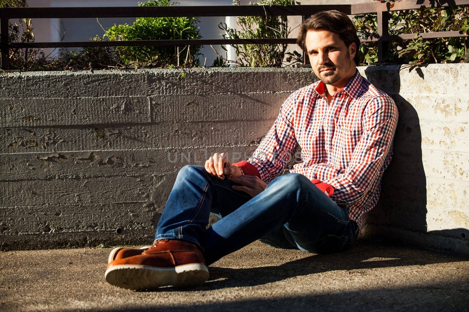 Young man with a checked shirt sits on a wall in the sun and relaxes
