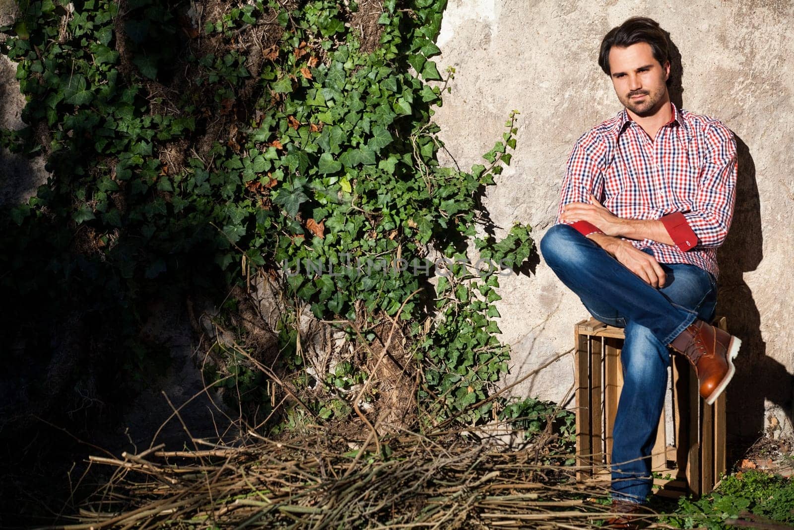 Young man with a plaid shirt and jeans with dark hair and beard outdoors under an efeuranke in daylight