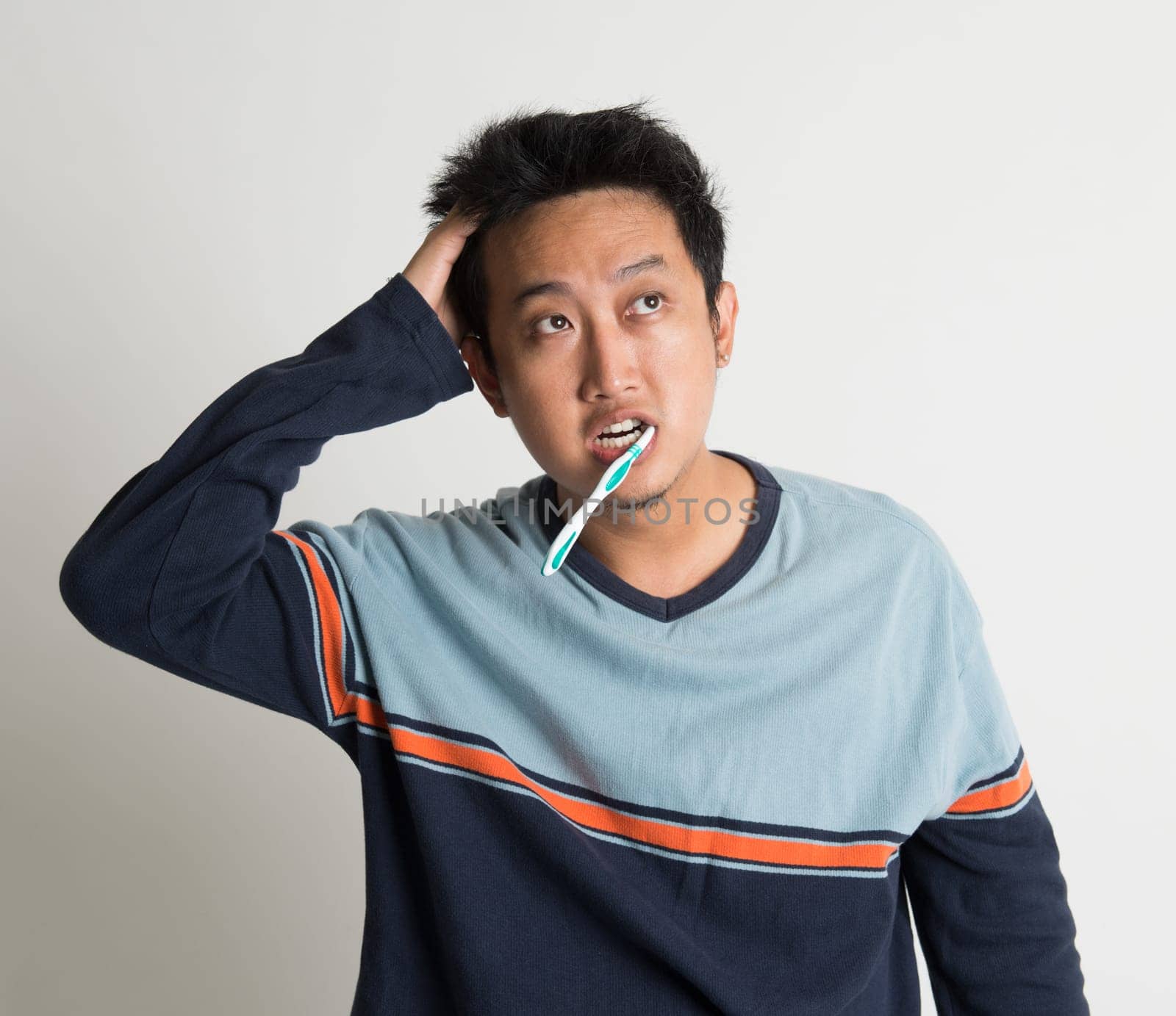 Southeast Asian male scratching head and looking up while brushing teeth in a morning, on plain background