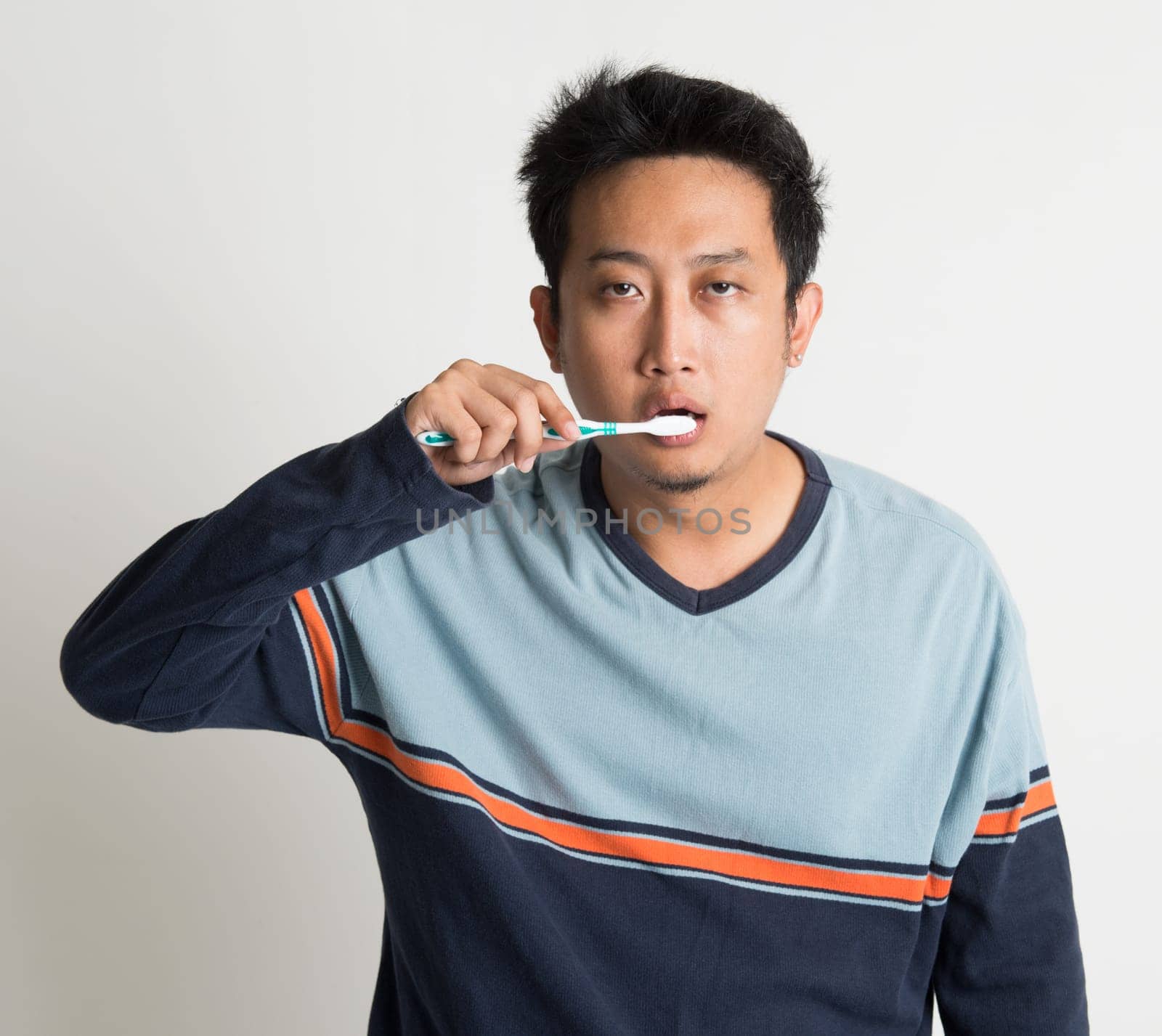 Southeast Asian male brushing teeth with sleepy eyes in a morning, on plain background