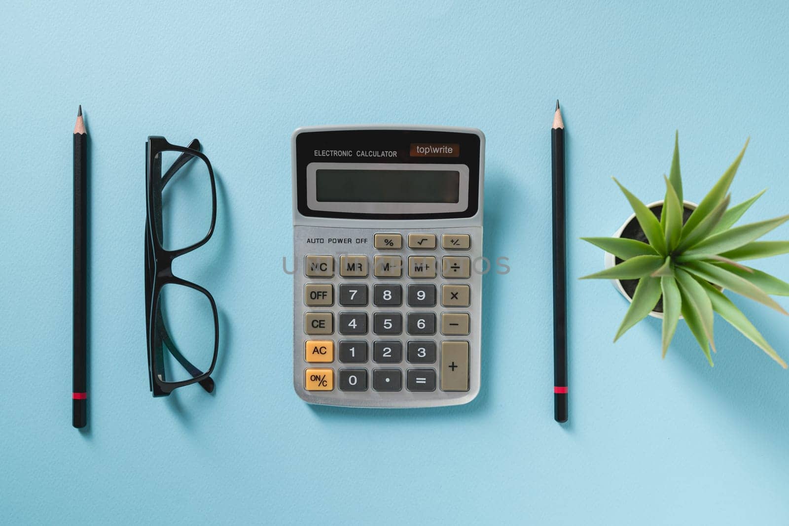 Top view of calculator, glasses, pen and green plant on blue office desk by Sonat