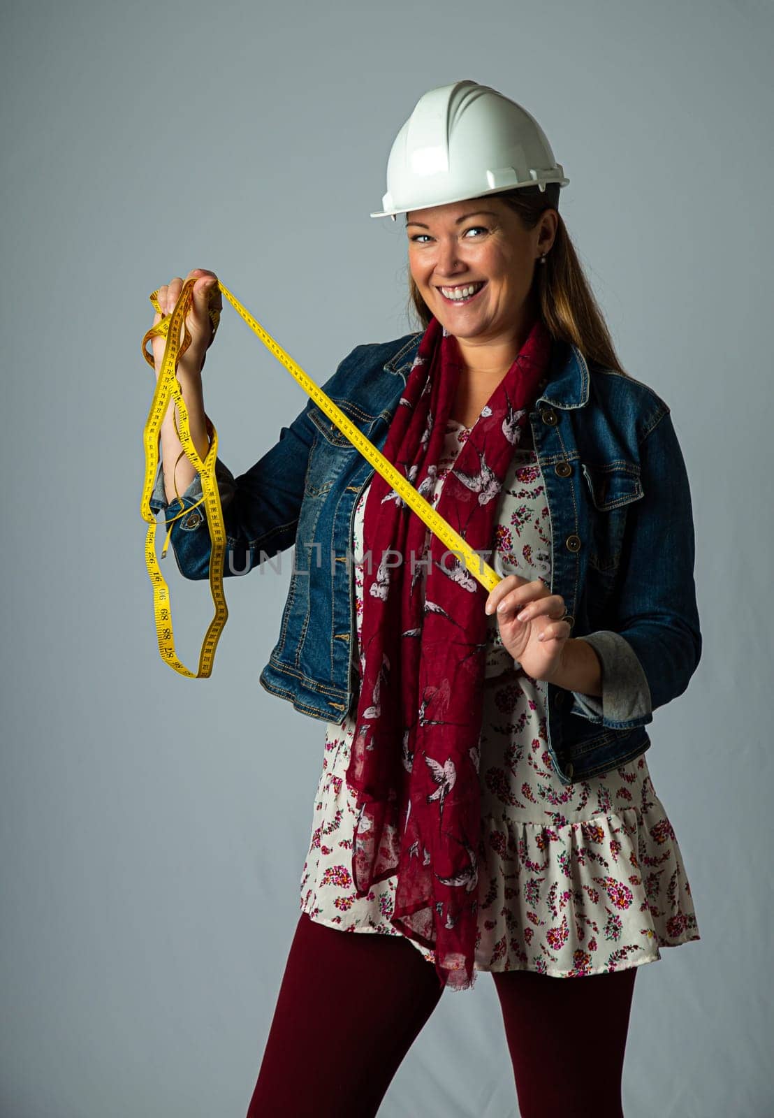 Forty something woman, wearing stylish bohemian clothing and a hard hat, holding a measuring yellow tape