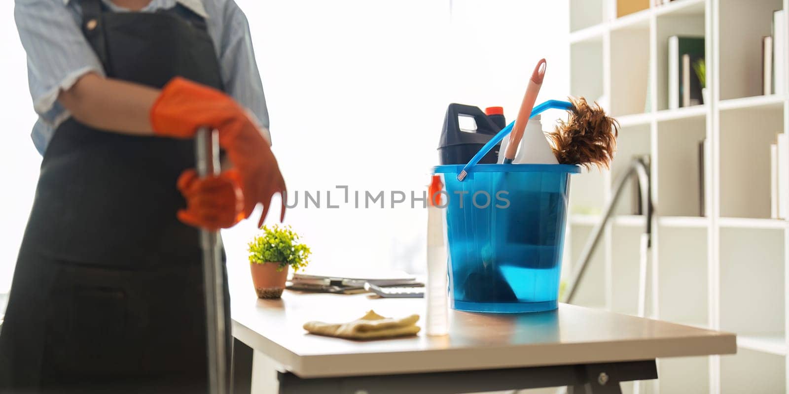 A cleaning woman is standing inside a building holding with a blue tank on the side facilities for tidying up in her hand.